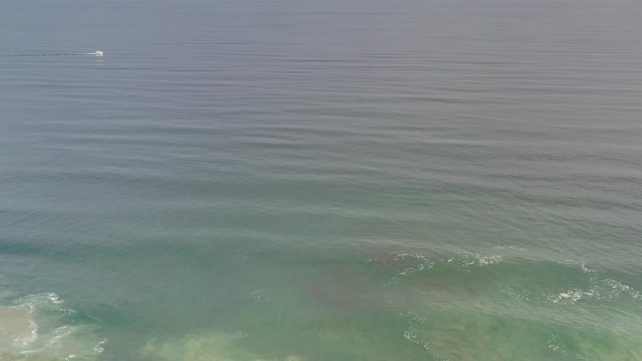 Los Cabos ocean view with a small white boat near the upper left, blue-gray water, green shallows, pale surf at the bottom edge, and no visible people.