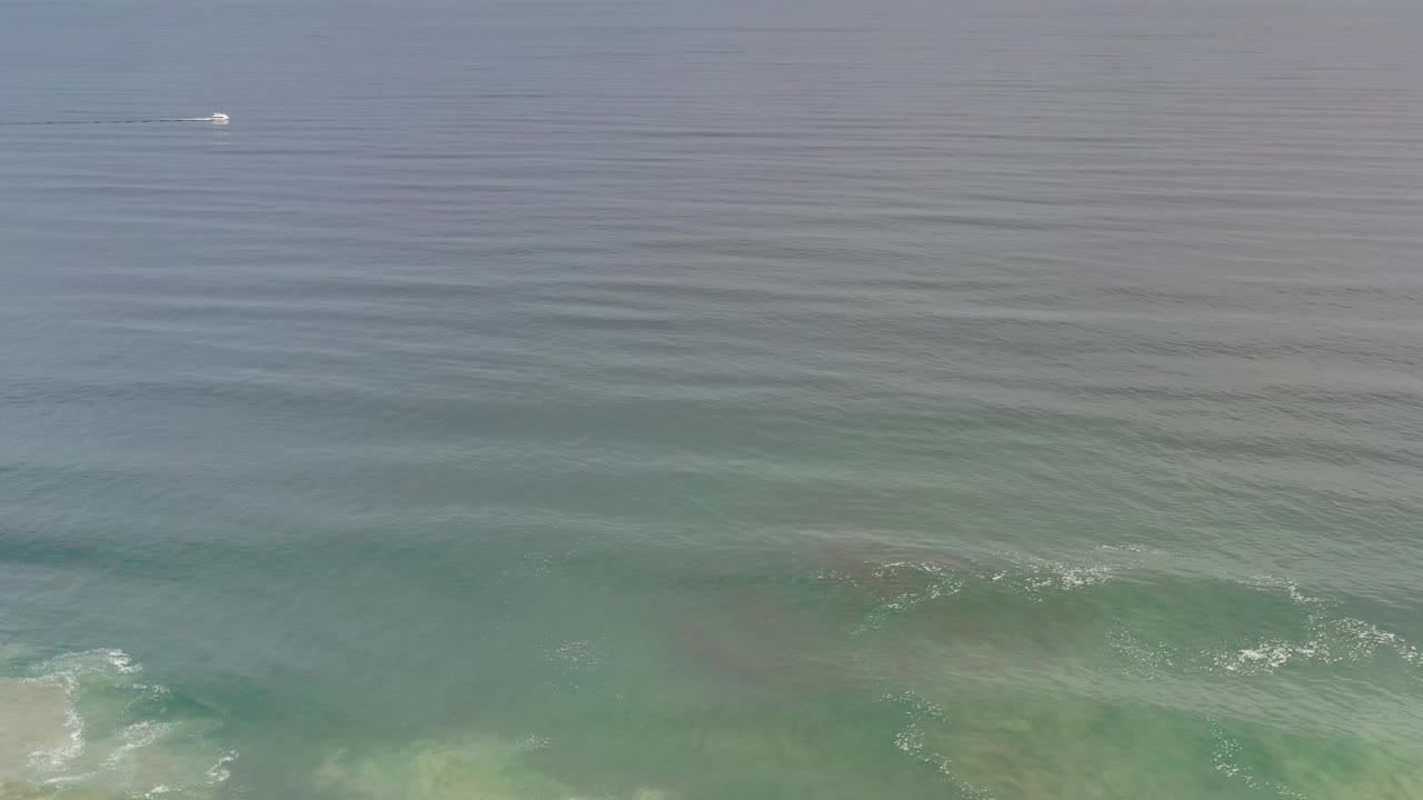 Los Cabos water fills the frame as a small white boat sits high in the image, with rippled blue surface texture and low white surf along the lower edge, no visible people.