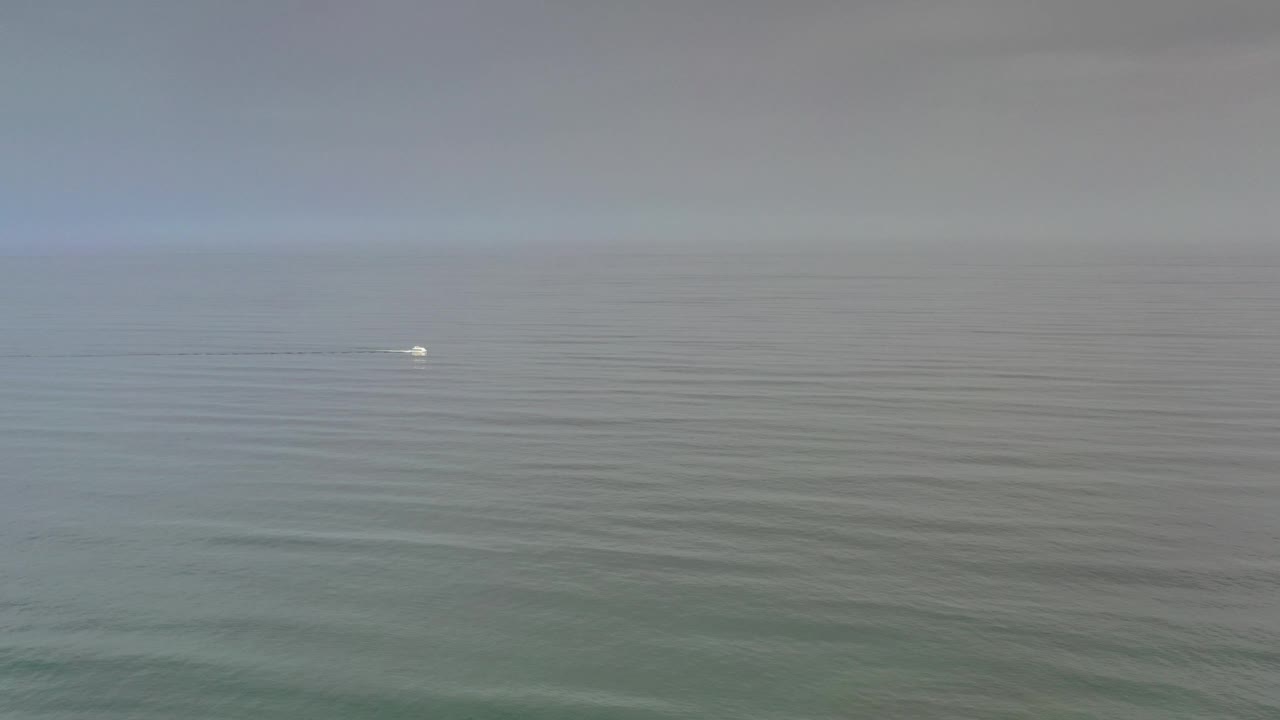 Los Cabos ocean seen in a wide elevated view with gray-green water, pale cloud cover, a faint white vessel or wake near the left side, and no visible people.