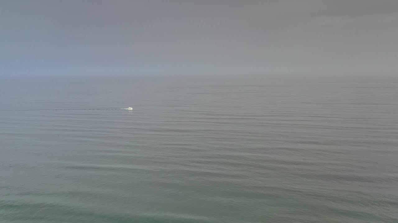 A wide Los Cabos sea view shows soft ripples, a hazy horizon, overcast sky, a small white offshore detail slightly right of the previous frame, and no visible people.