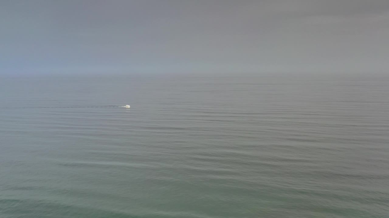 Muted ocean surface below a pale cloudy sky with a tiny white wake.