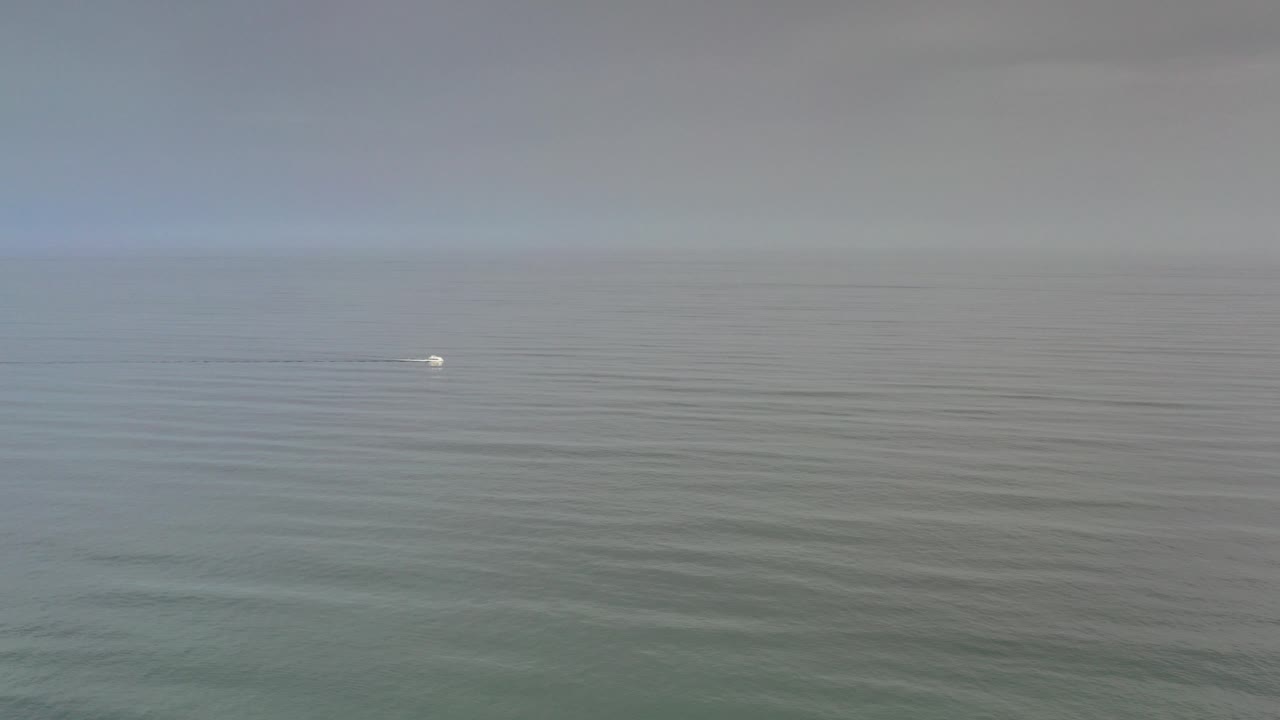 Wide Los Cabos ocean view at the start of the clip, with a hazy horizon, muted blue-gray water, a tiny white circular mark near center, and no visible people.