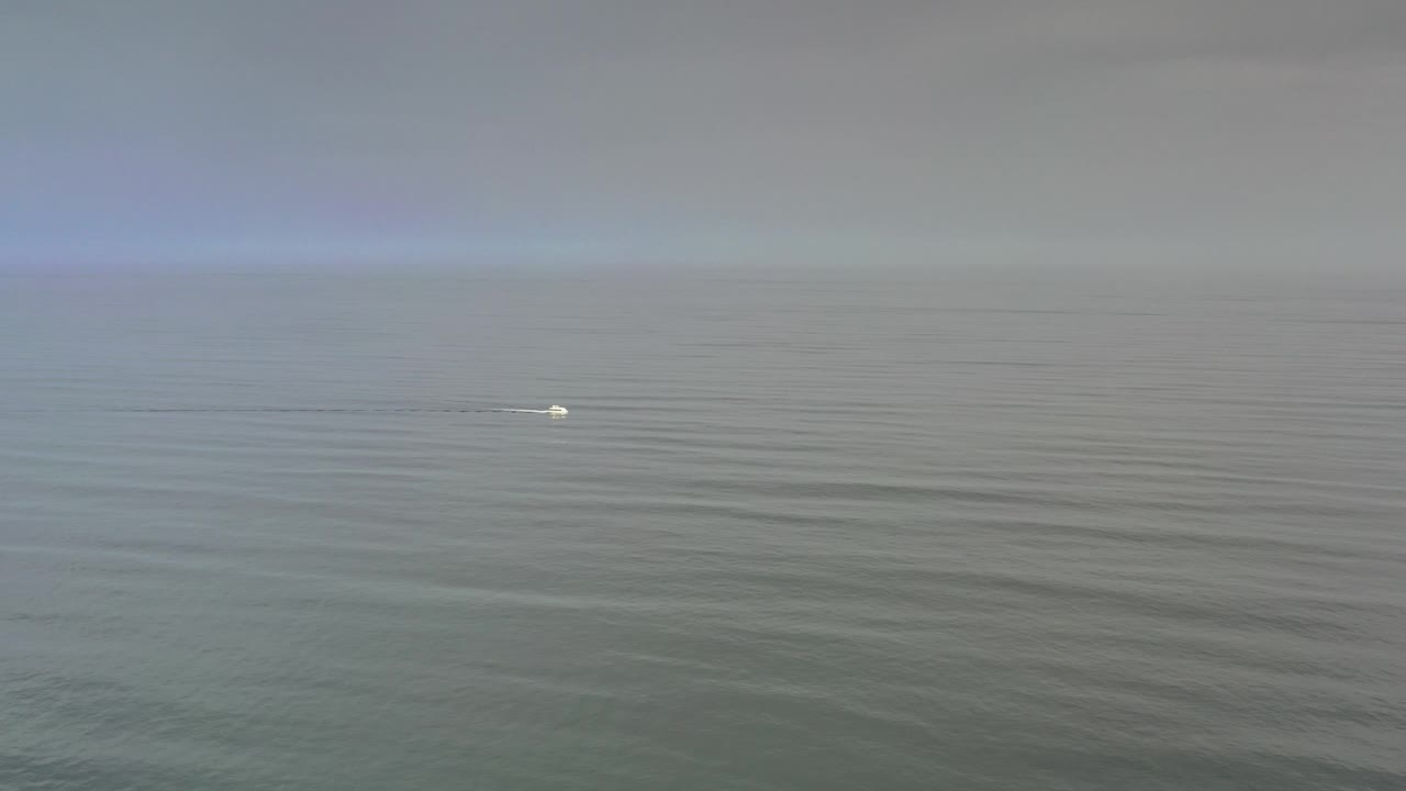 Tiny white boat with wake on open water below an overcast horizon.