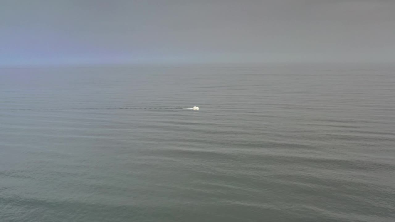 The Los Cabos water remains calm under a hazy cloud layer, with the small white object near the middle distance and no visible people.