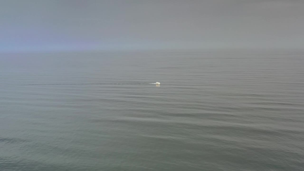 Fog-muted Los Cabos ocean view with low ripples across the foreground, a pale horizon, a small white boat or wake nearer the middle of the frame, and no visible people.