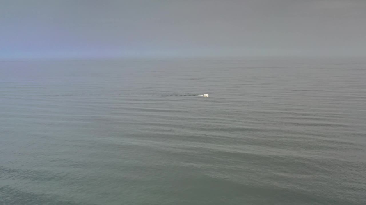 At 0:01, the distant boat appears slightly farther across the open Los Cabos water, with soft ripples below a pale hazy sky and no visible people.