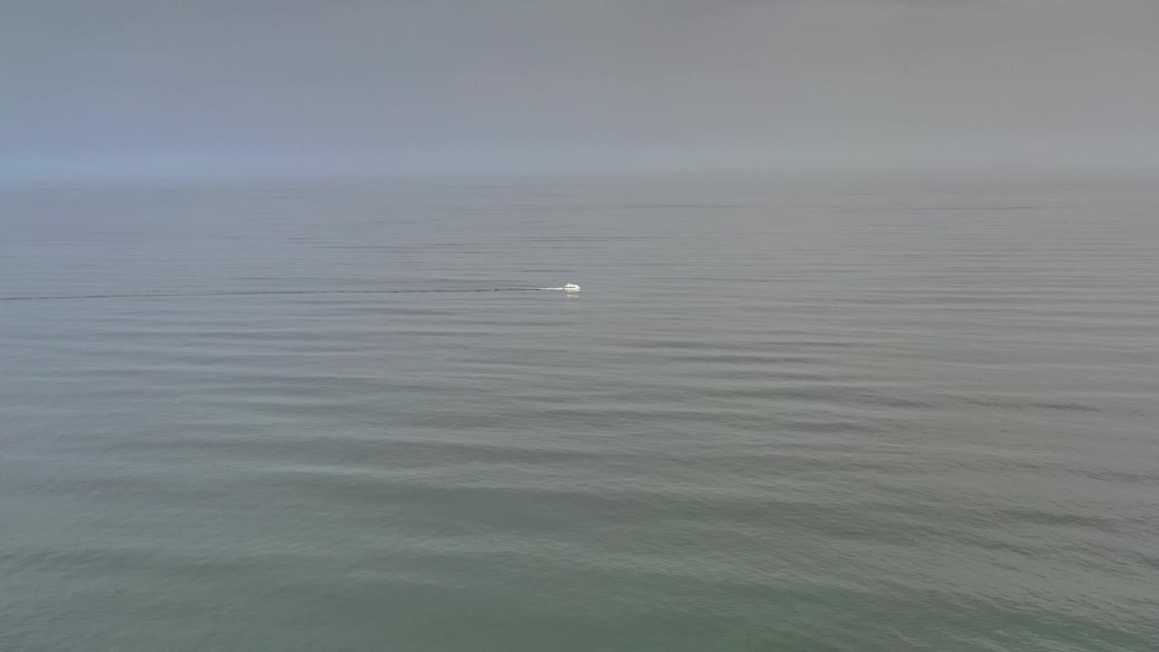 Los Cabos open water frame with the white boat slightly farther right, a thin wake trailing left, muted gray blue water, and no visible people.