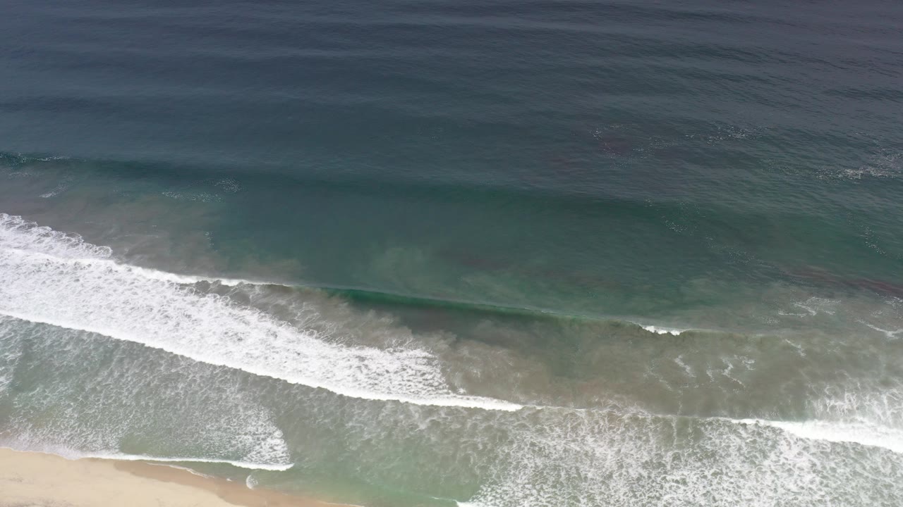 Overhead view of teal water and white surf reaching a sandy Los Cabos beach, with no visible people.