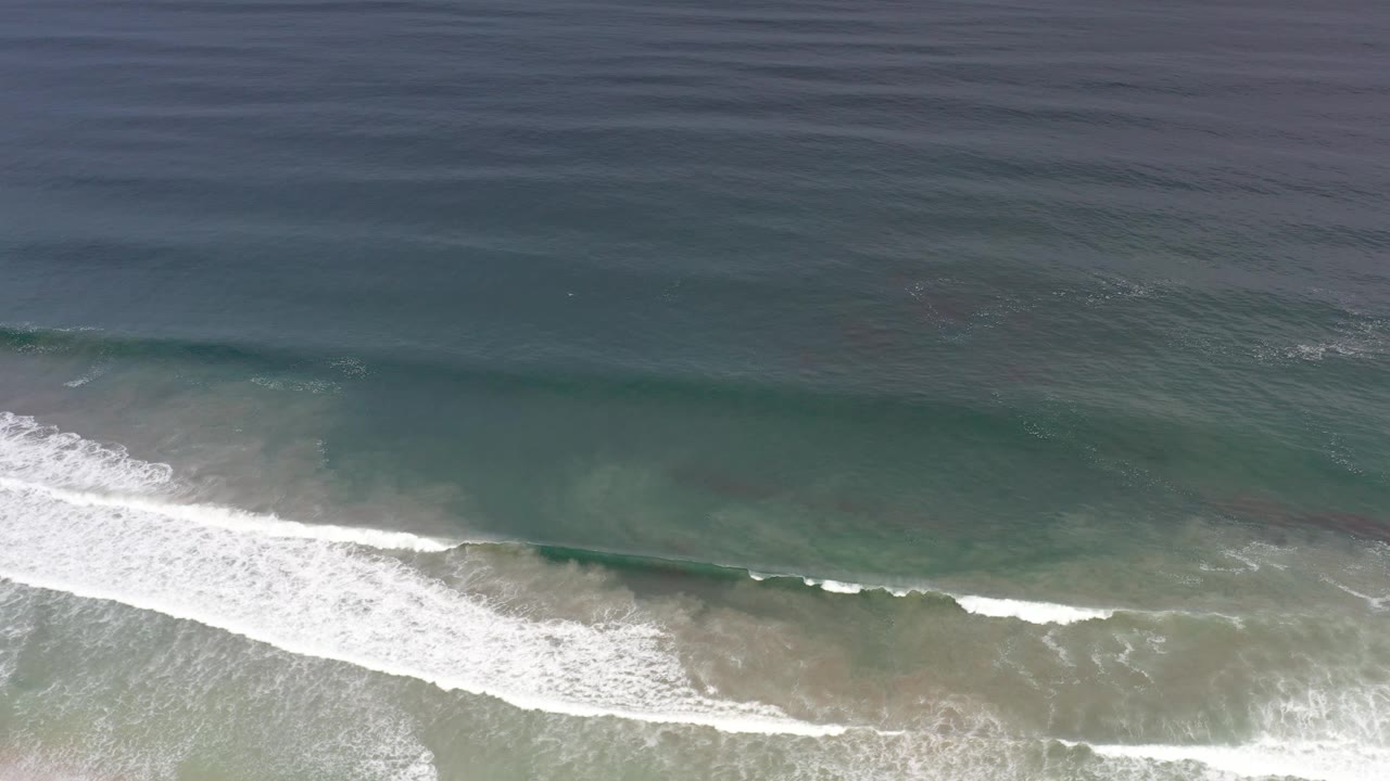 Aerial view of darker offshore water above a breaking surf band along a sandy Los Cabos beach, with no visible people.
