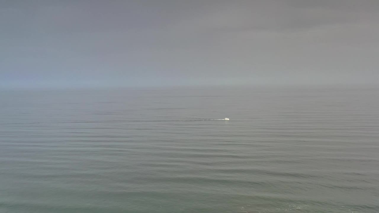 Los Cabos water and horizon remain wide in frame, with the small white offshore point slightly right of center and no visible people.