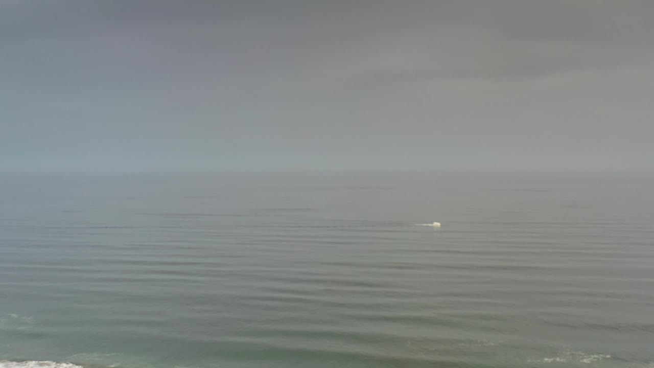 A later Los Cabos frame holds on the broad sea, with the small white point slightly right of center and thin horizontal swell bands across the water, no visible people.