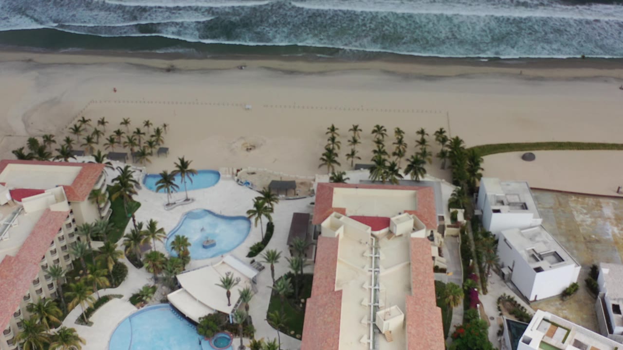 Overhead view of beachfront buildings, pools, palms, sand, and surf.