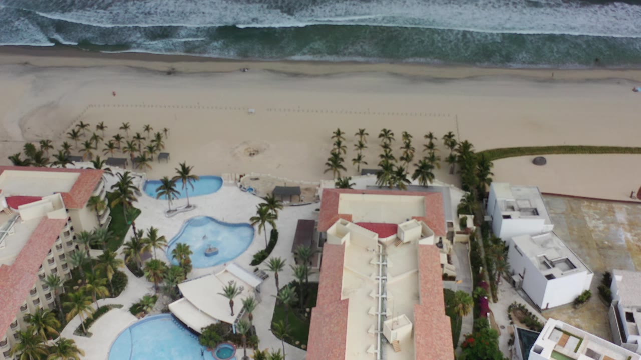 Overhead view of hotel pools, palm trees, sandy beach, and breaking waves.
