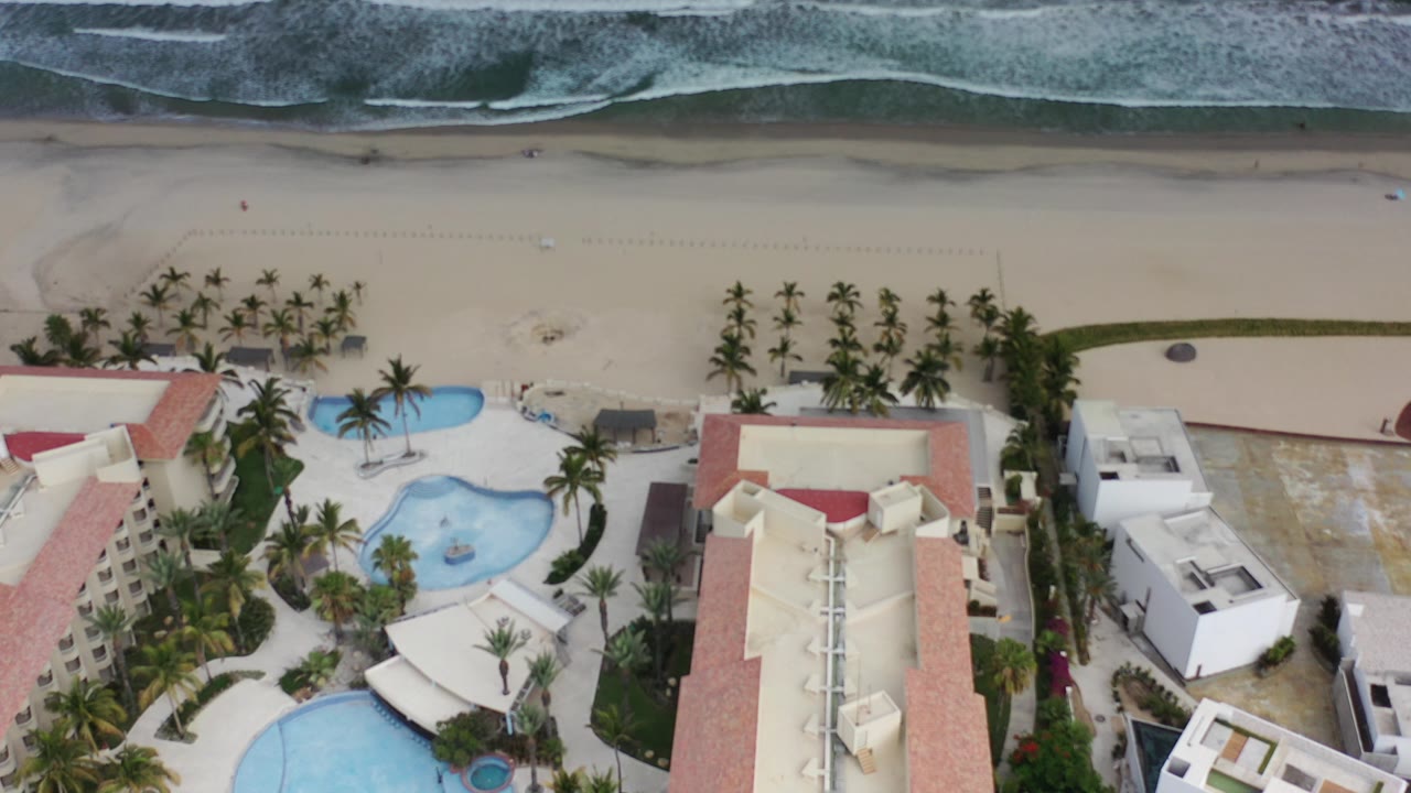 Aerial view of a beachfront hotel, pools, palms, sand, and surf.