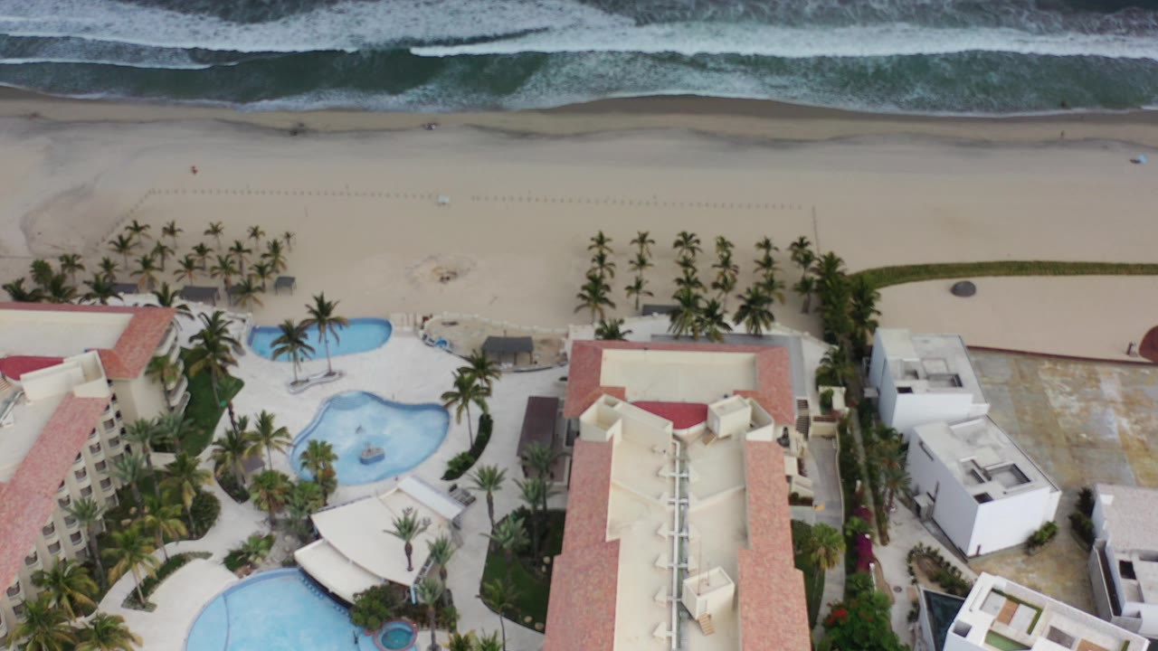 High aerial frame over Los Cabos beachfront buildings with red roofs, blue swimming pools, palm rows, wide sand, ocean surf, and no visible people.