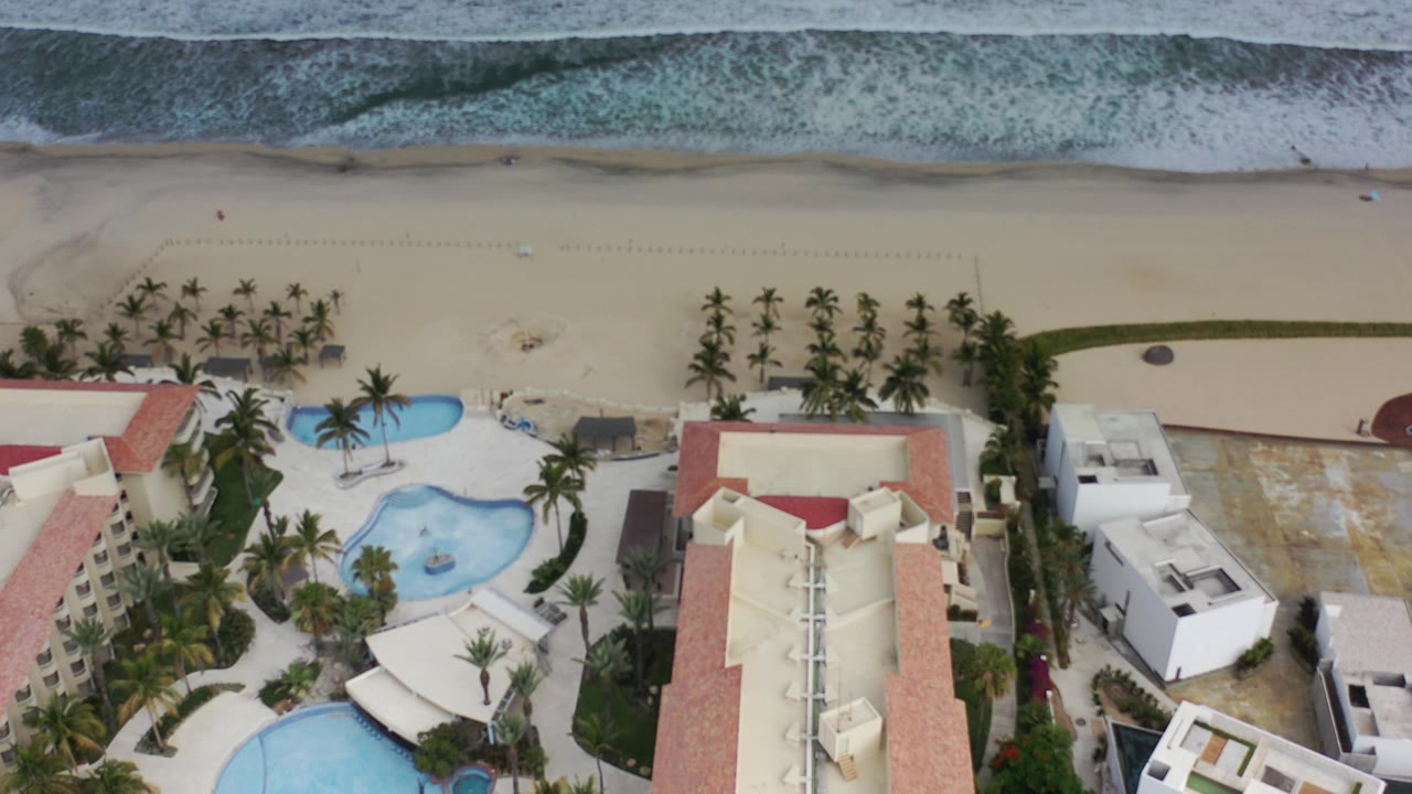 High aerial view in Los Cabos, Mexico showing red-roof resort buildings, curved blue pools, palm trees, pale beach sand, and a foamy surf line, with no visible people.