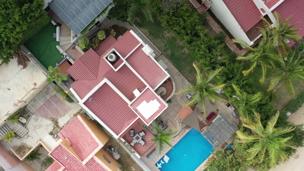 Overhead frame from Los Cabos, Mexico showing a red-tile house roof with white trim, palms, paved patios, shrubs, and a blue pool at the lower right, with no visible people.