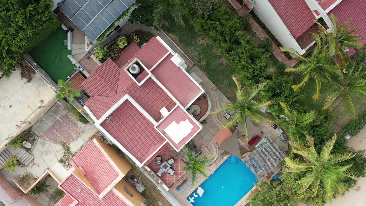 Top-down Los Cabos, Mexico view with the red roof slightly larger in frame, palm crowns along the right side, paved walkways, shrubs, and the pool corner visible, with no visible people.