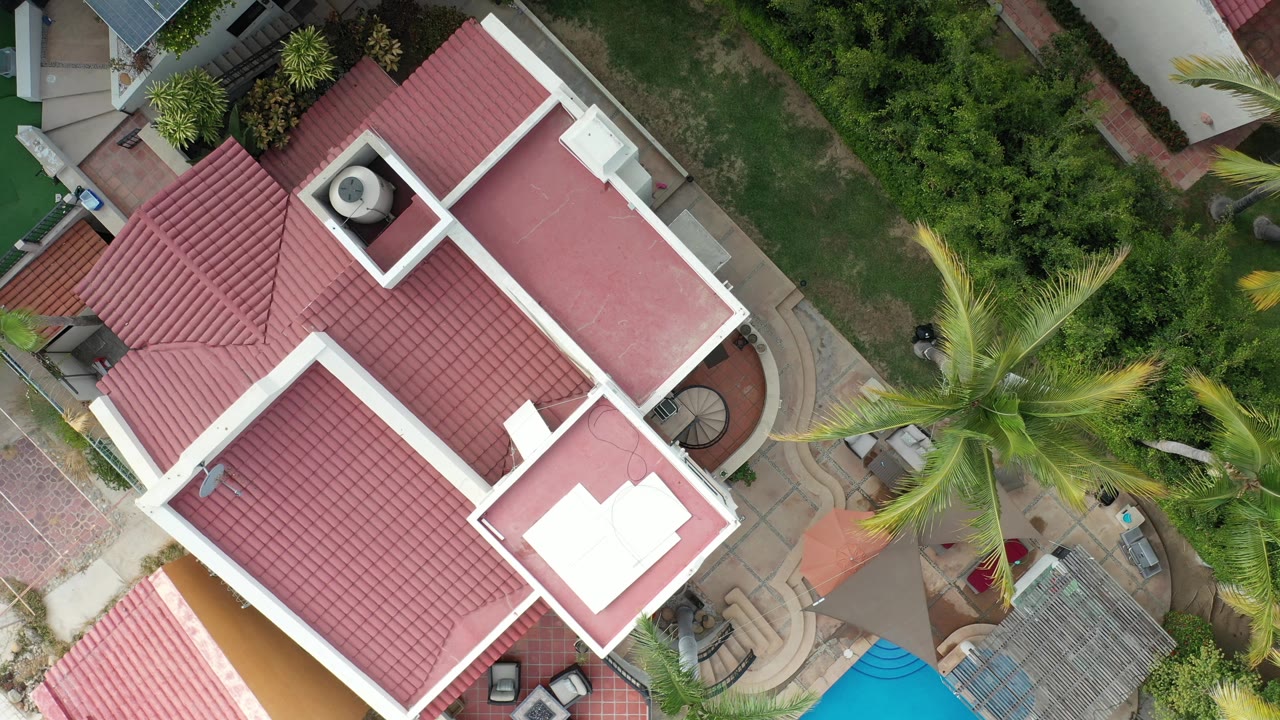 Los Cabos overhead frame with the red roof shifted slightly in view, a central palm tree, curved patio paths, green lawn, shrubs, and no visible people.