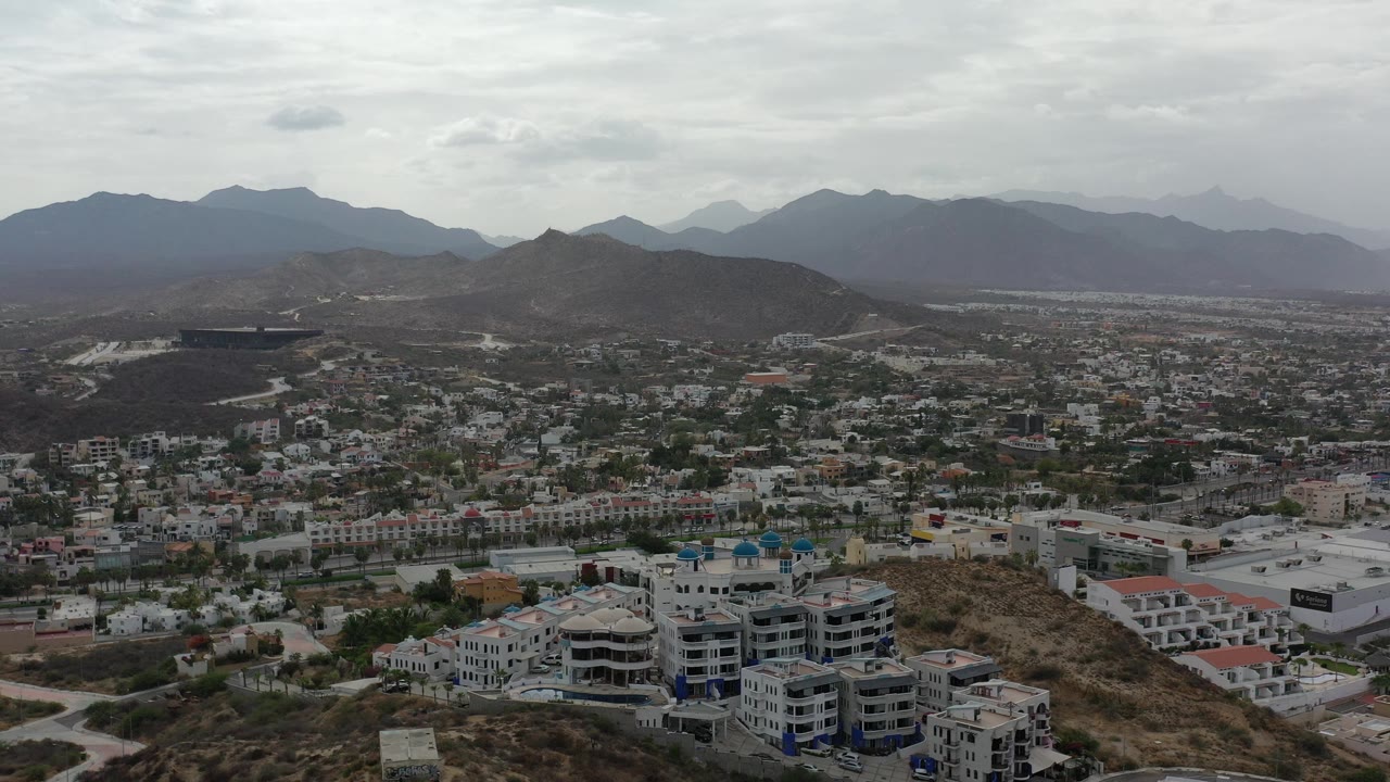 Aerial view of apartment buildings, urban blocks, dry hills, and distant mountains.
