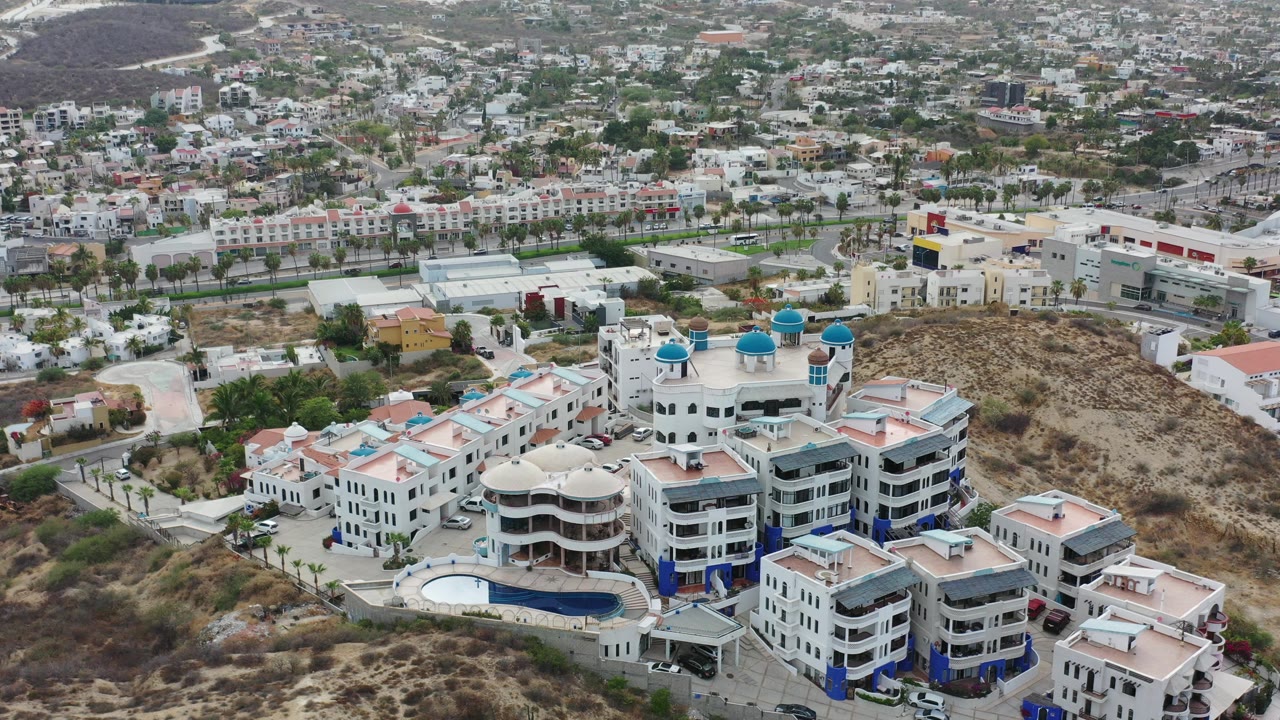 High aerial view in Los Cabos, Mexico, showing white resort buildings with blue domes, a curved pool, dry hillside, roads, and residential blocks, with no visible people.