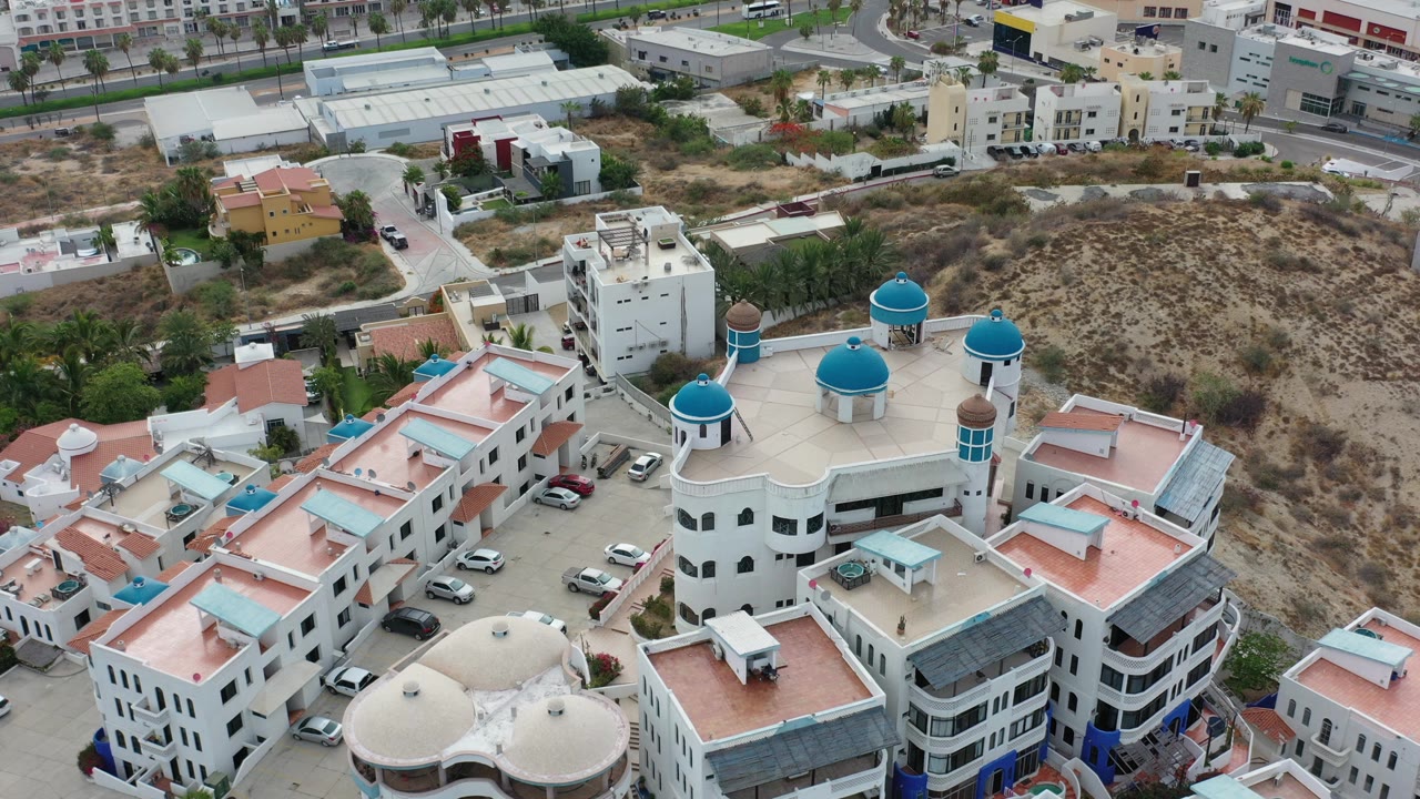 High aerial view in Los Cabos, Mexico showing a white building complex with blue domes, terracotta roofs, parked cars, dry lots, and no clearly visible people in this opening frame.
