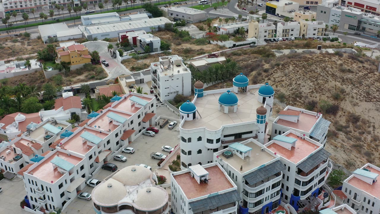 Oblique drone frame over Los Cabos, Mexico with blue-domed rooftops in the lower right, parked cars near the buildings, dry brushy land behind, and no clearly visible people.