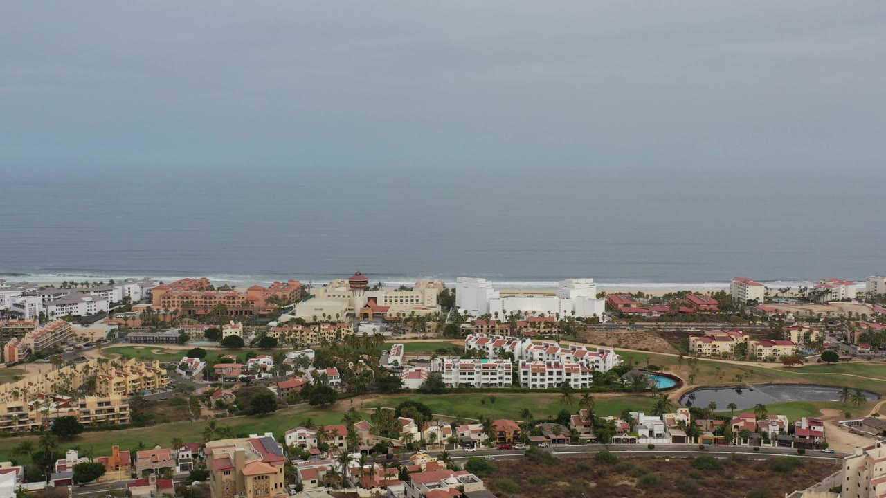 Opening aerial frame over Los Cabos, Mexico, with low-rise buildings, green grounds, a pale surf line, blue-gray ocean, hazy sky, and no visible people.