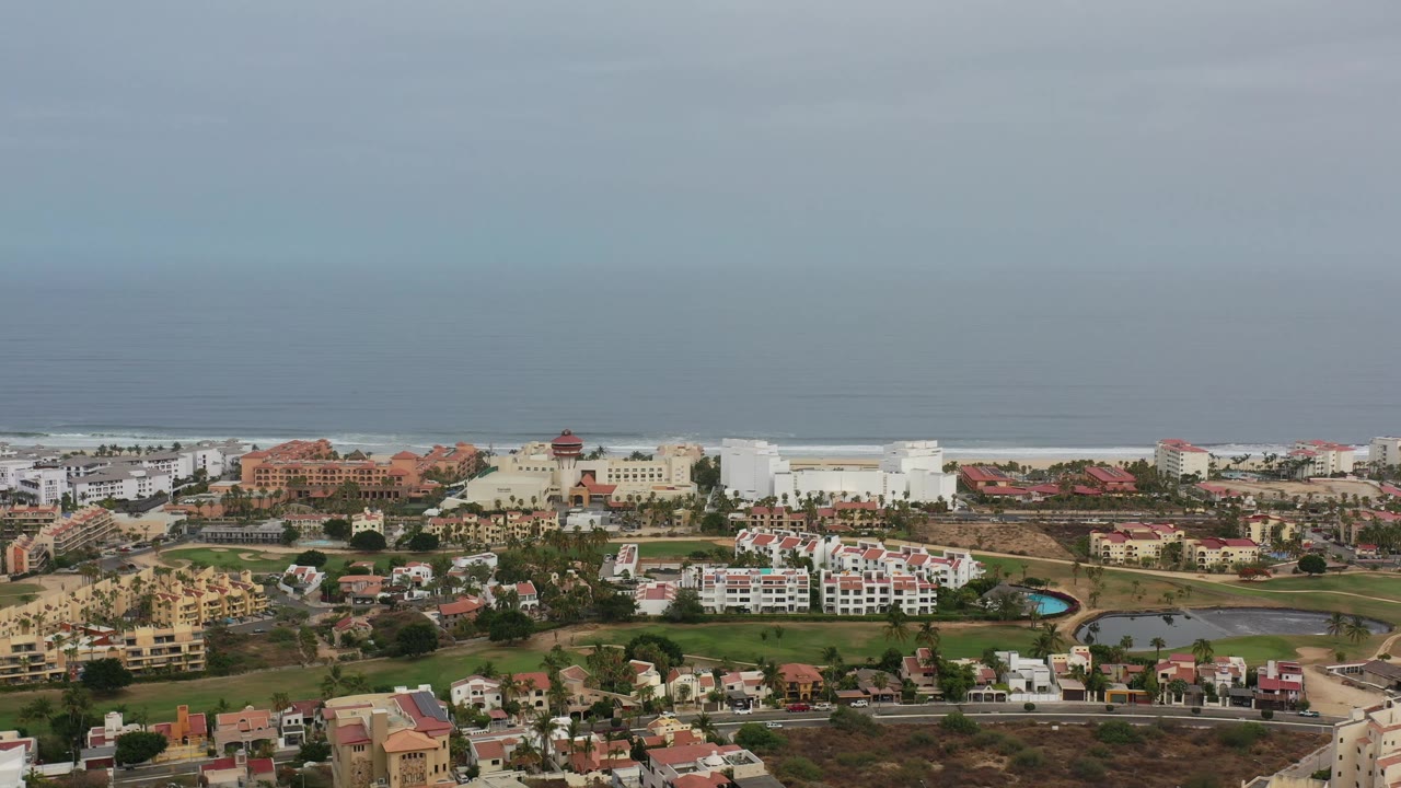 Early aerial frame of Los Cabos showing clustered homes, larger coastal buildings, green landscaping, distant ocean, muted daylight, and no visible people.