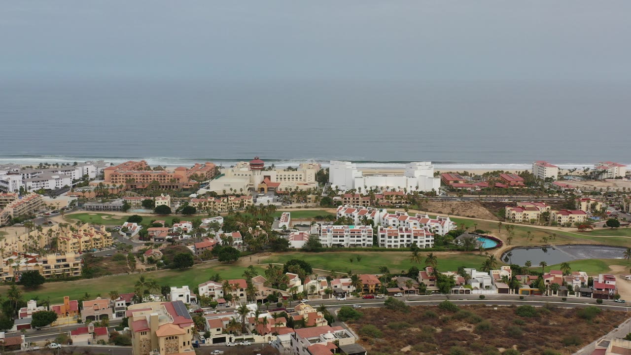 Opening aerial frame over Los Cabos, Mexico, showing clustered homes, larger pale seaside buildings, green lawns, roads, and a hazy ocean horizon with no visible people.