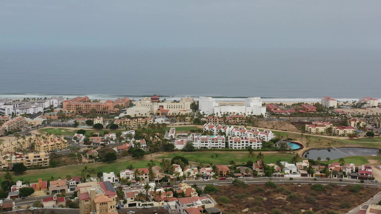 Aerial frame over Los Cabos with red-roofed homes in the foreground, a green landscaped area through the middle, pale buildings near the shore, and no visible people.