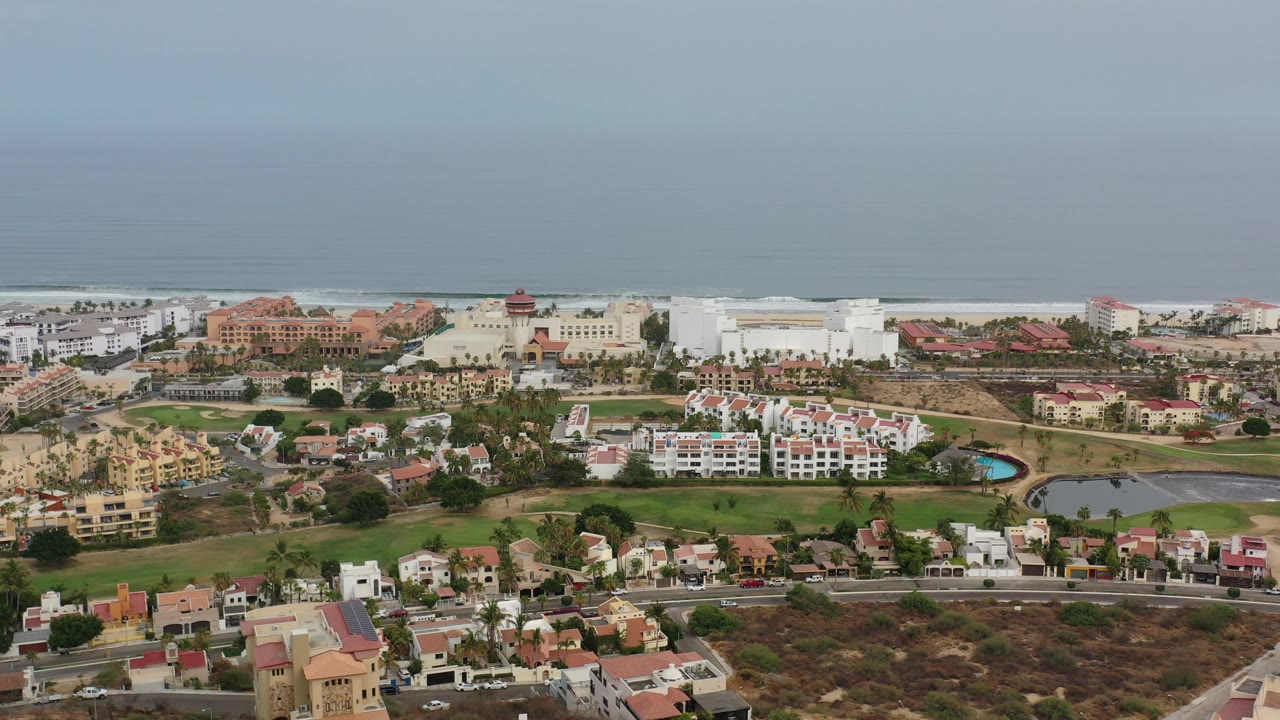 Wide aerial of seaside buildings, houses, roads, green areas, and ocean backdrop.