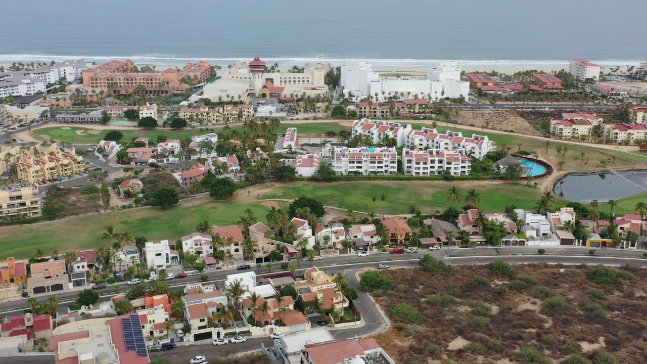 Opening aerial frame over a Los Cabos residential area with terracotta-roof homes, green lawns, curving roads, large coastal buildings, ocean surf in the background, and no visible people.