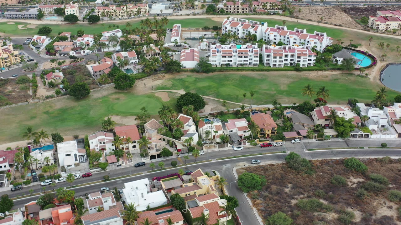 Aerial view in Los Cabos, Mexico showing residential blocks, a road with parked vehicles, green landscaping, a small turquoise pool, and no visible people.