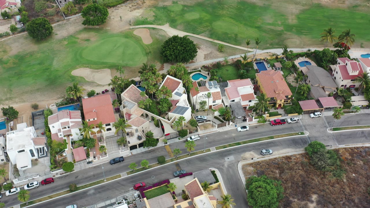 High aerial view over Los Cabos homes with red and white roofs, pools, palm trees, a road, parked cars, golf-course grass, and no visible people.