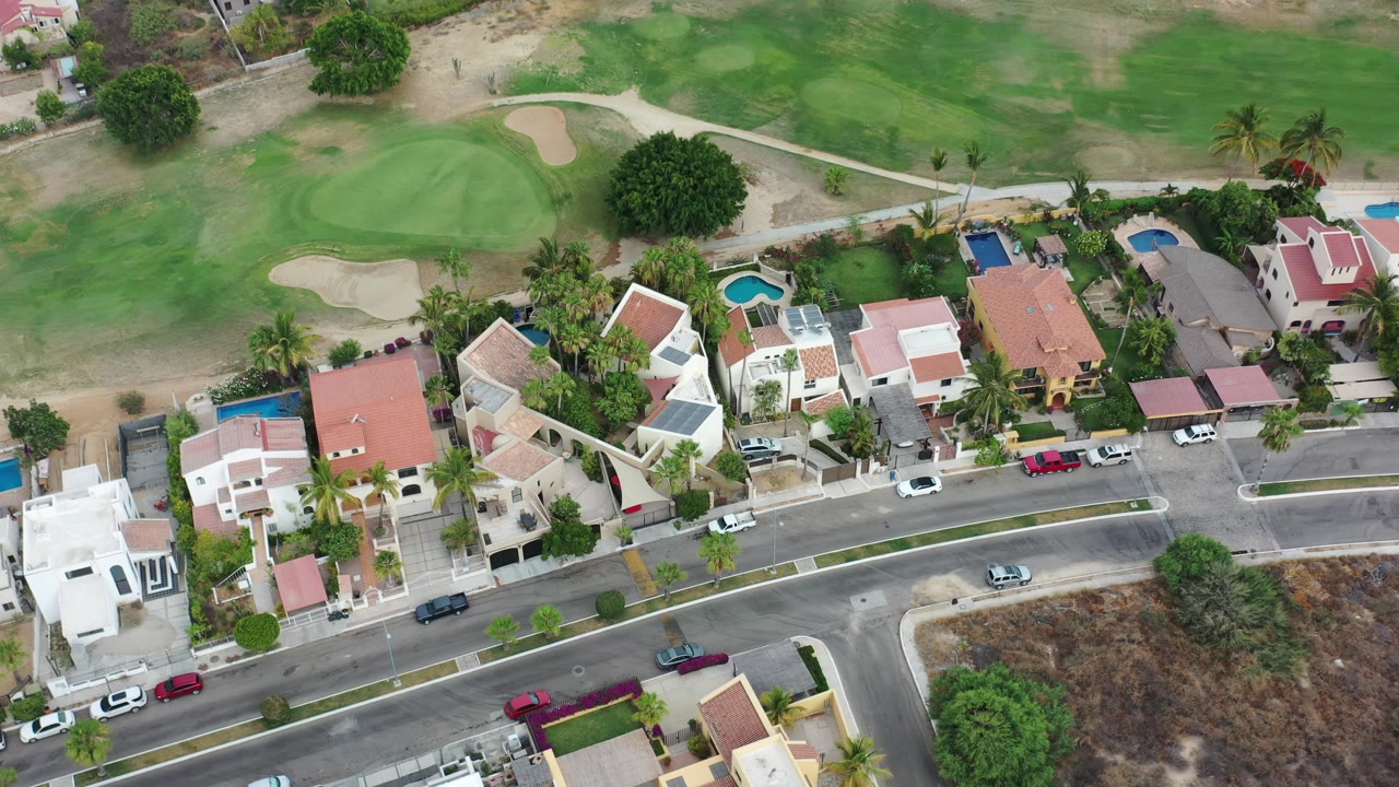 Aerial frame in Los Cabos showing a row of private houses, small blue pools, parked vehicles, palm trees, a curved street, golf-course turf, and no visible people.