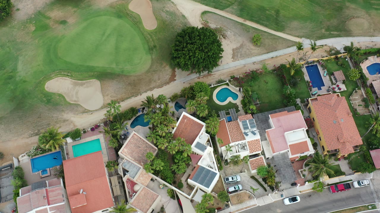 Overhead aerial frame in Los Cabos, Mexico showing modern houses with private pools along a road beside a golf green and sand bunker, with no visible people.