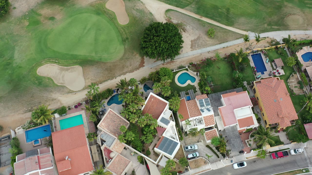 A Los Cabos overhead view shows tile-roof homes, blue swimming pools, parked cars, palms, a golf green, dry turf, and no visible people.