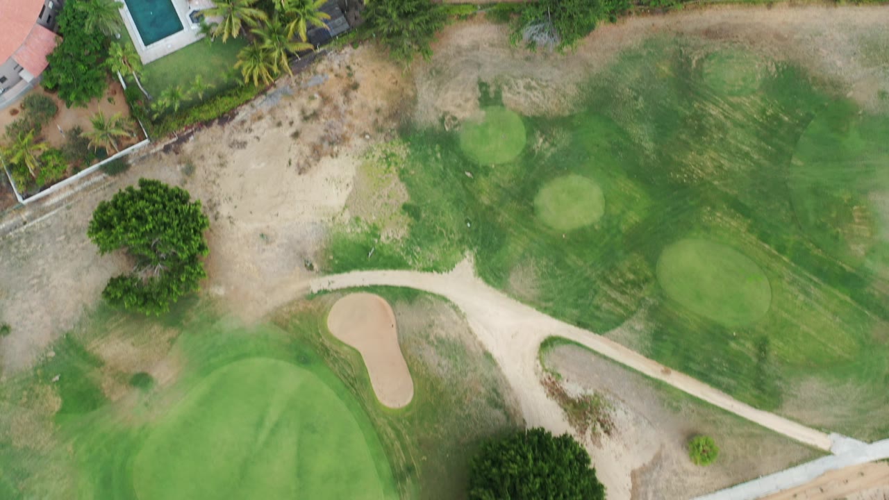 Overhead view of golf course grass, sand path, palm trees, and nearby homes.