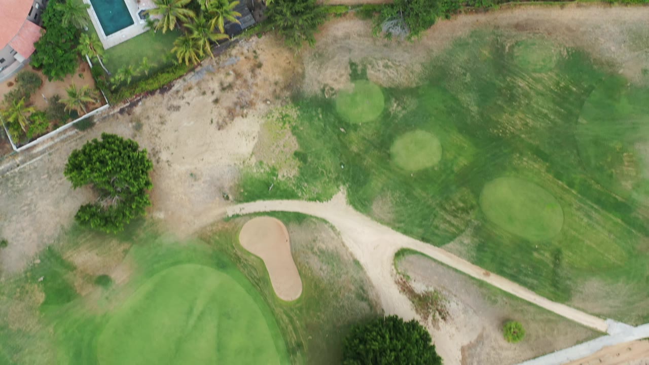 Overhead Los Cabos frame with a fairway, rounded greens, a sand trap, dry rough, palm trees, property walls, and no clearly visible people.
