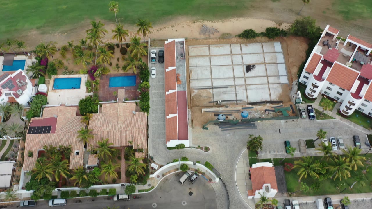 Aerial Los Cabos frame with the golf course at top, a road through the middle, low-rise residences, blue pools, palms, and no clearly visible people.