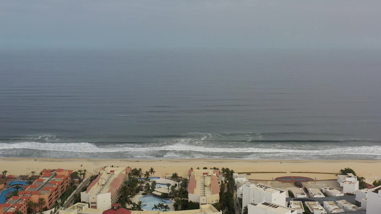 Los Cabos shoreline view with long horizontal wave lines approaching the sand and low beachfront buildings partly visible at the lower edge, no visible people.