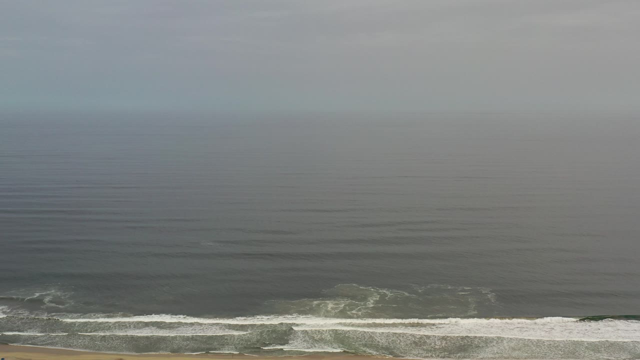 Aerial view of gray-blue ocean surf meeting a narrow sandy beach.