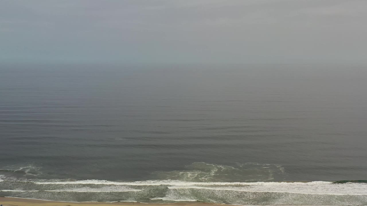 Aerial Los Cabos shoreline frame with darker water across the upper area, a thin white wave line near shore, tan beach below, and no visible people.