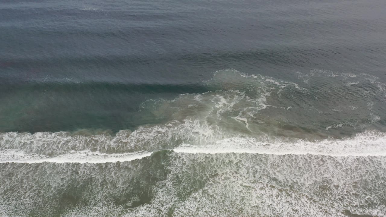 Top-down aerial frame over Los Cabos, Mexico, showing green ocean water, a dark wave band, and white surf foam spreading along the lower edge with no visible people.