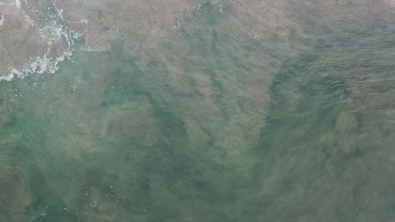 Overhead Los Cabos water shows darker sand or mud patches under the green surface, with broken white foam near the top and no visible people.