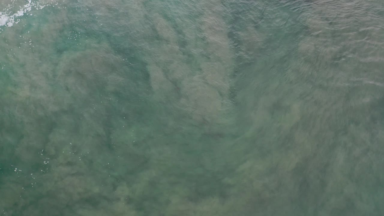 Overhead view of teal shallow water in Los Cabos, Mexico, with loose sand or mud visible below the surface and no visible people.
