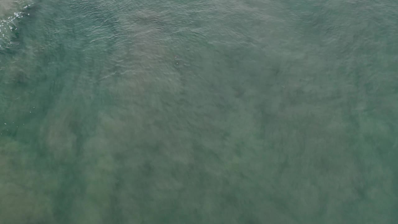 Aerial frame of shallow Los Cabos ocean with a diagonal line of small white wave foam crossing green water and no visible people.