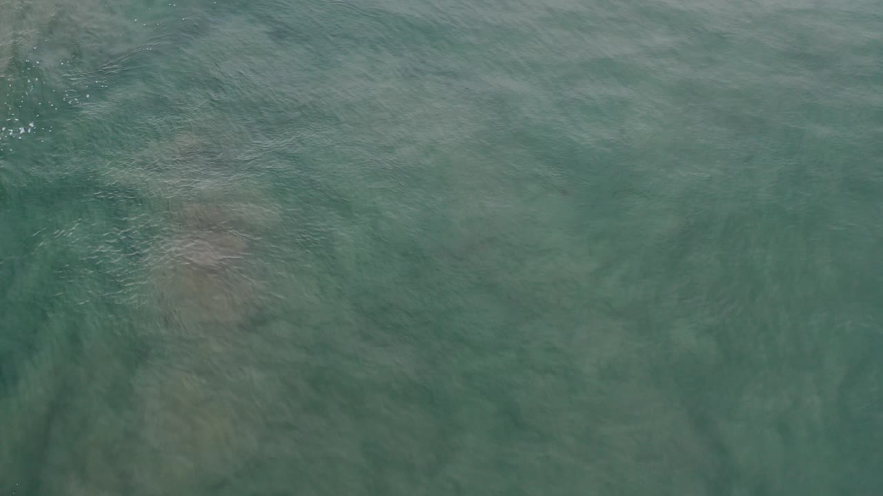 Overhead view of green-blue water in Los Cabos, Mexico, with soft ripples and muted submerged patches, no visible people.