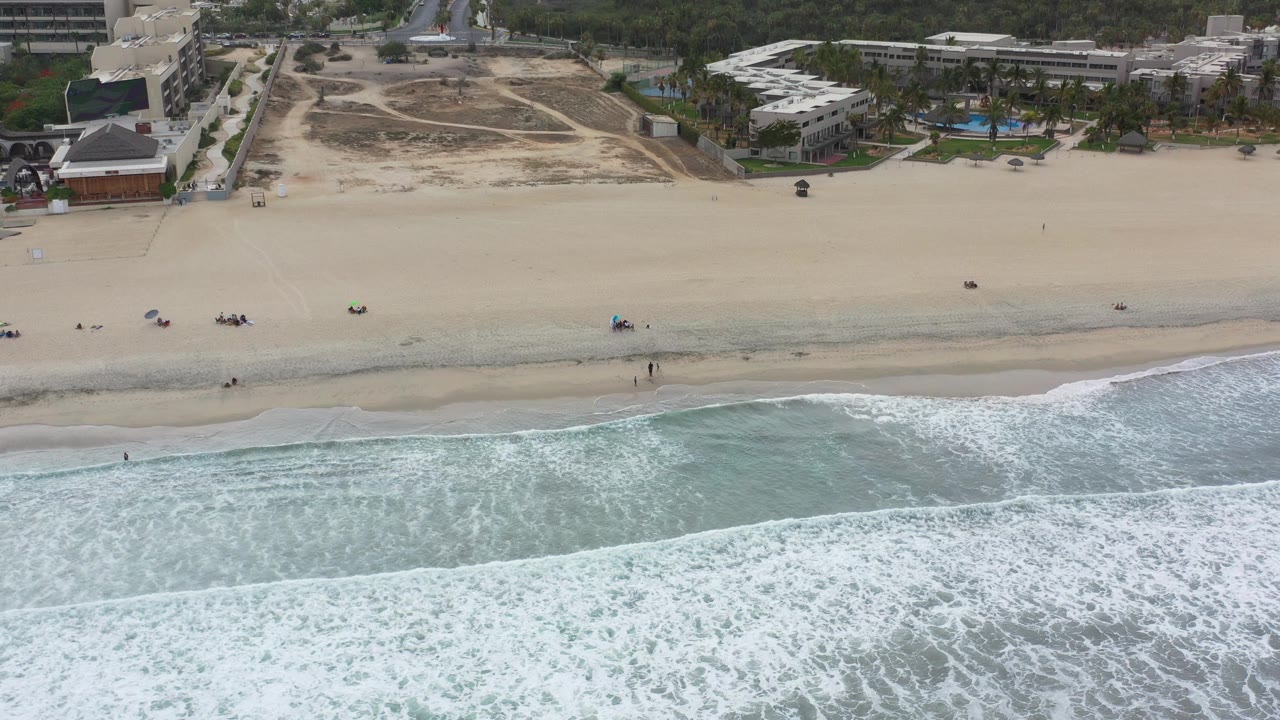 Aerial view in Los Cabos, Mexico, with white surf in the foreground, pale sand across the center, beachfront buildings beyond, and no clearly identifiable people.
